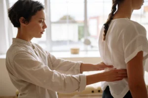 A healthcare professional examining a patient's lower back by gently pressing along the spine in a bright, sunlit room.